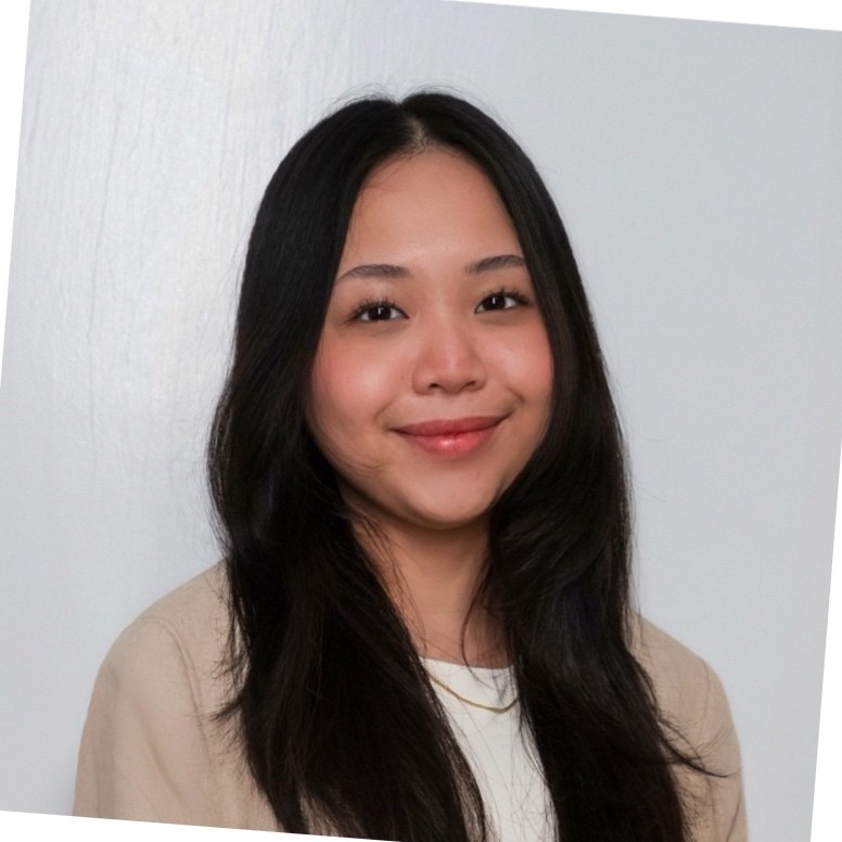 Female scientist with long brown hair. She has a slight smile, and is wearing a white shirt with a brown button up shirt over it.